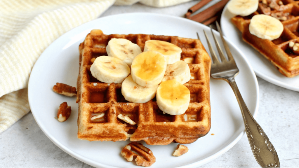 Golden-brown waffle topped with banana slices and pecan nuts on a white plate with a fork.